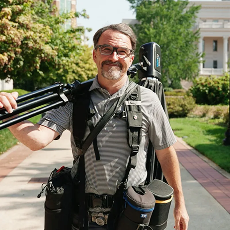 Belmont Photographer Sam Simpkins standing outdoors on the Belmont University campus, carrying camera equipment including a tripod and bags.