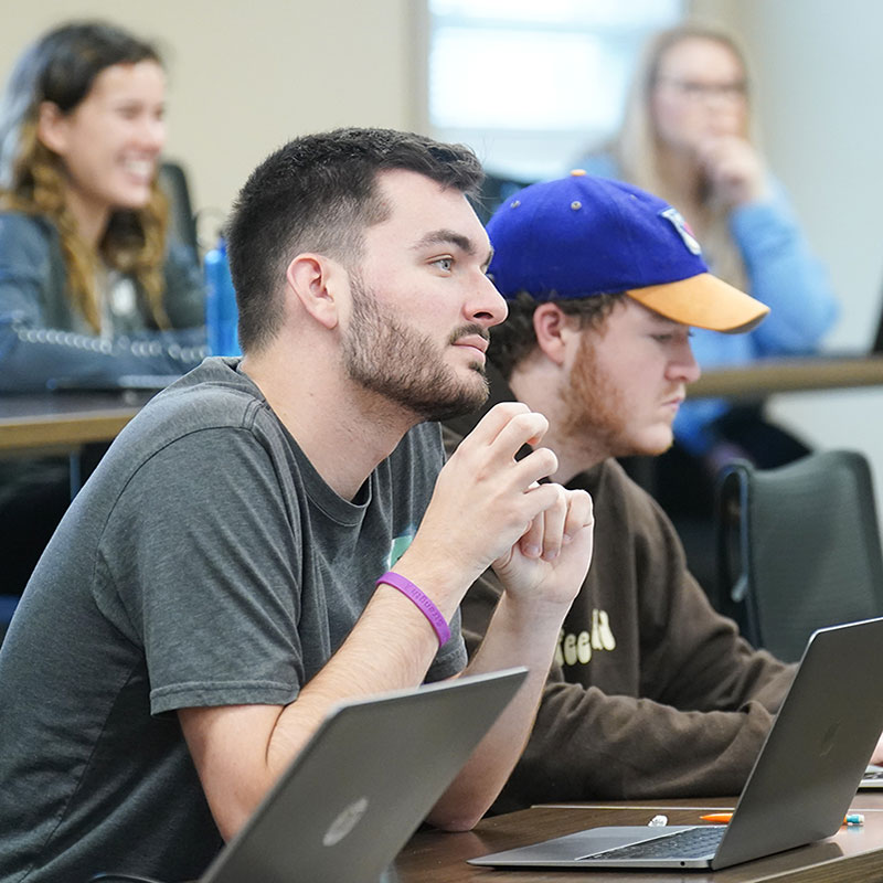 Two Male students sit in front of their laptops and look up, listening to a professor.