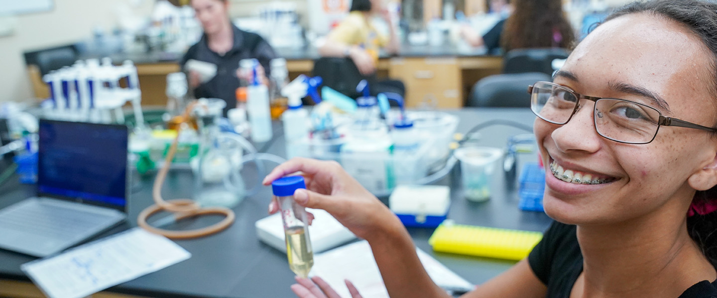 A female student holds up a test tube and smiles at the camera.