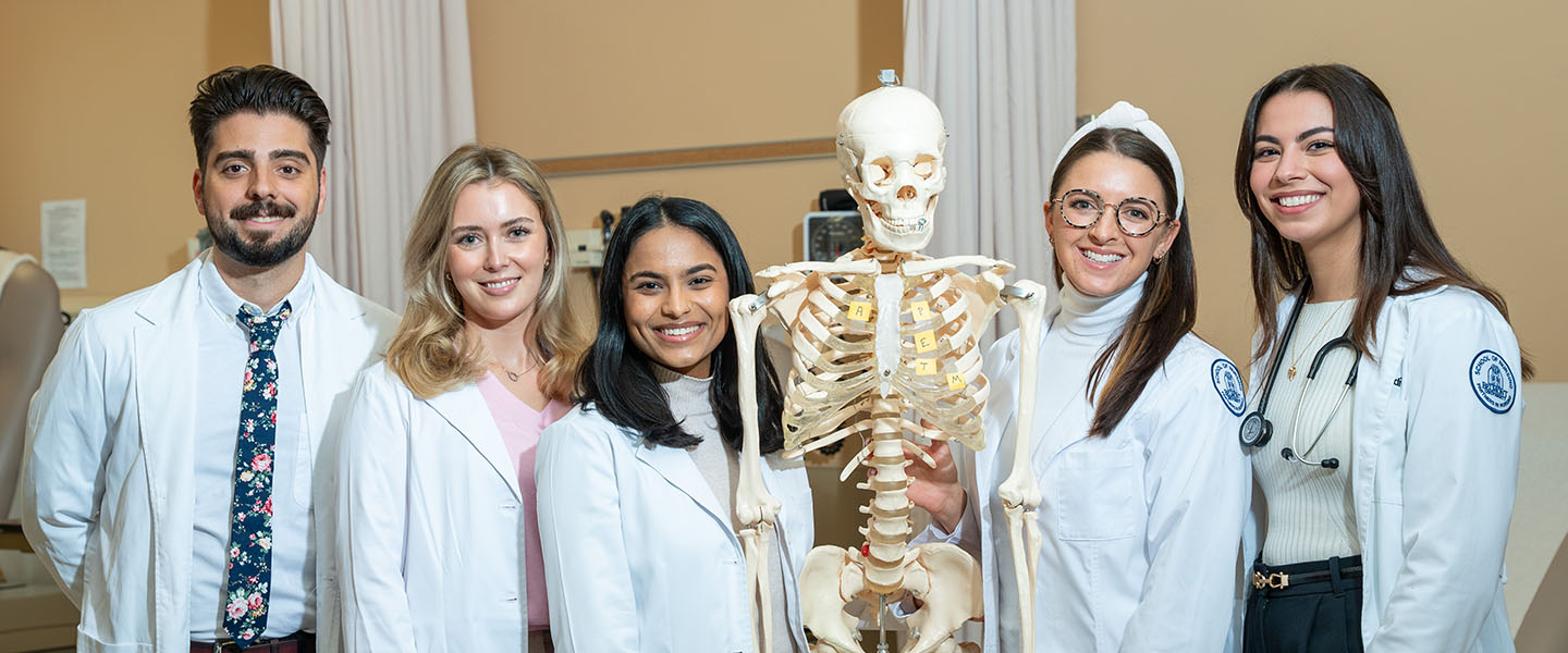 Five healthcare students, dressed in white lab coats, stand together in a clinical setting, smiling for the camera. They are positioned around a medical skeleton model used for study, which is placed prominently in the center. The group, diverse in appearance, exudes confidence and enthusiasm as they engage in their medical education. One student is holding the skeleton model, and another wears a stethoscope around her neck, indicating their healthcare focus. The background features medical equipment, reinforcing the professional, academic environment.