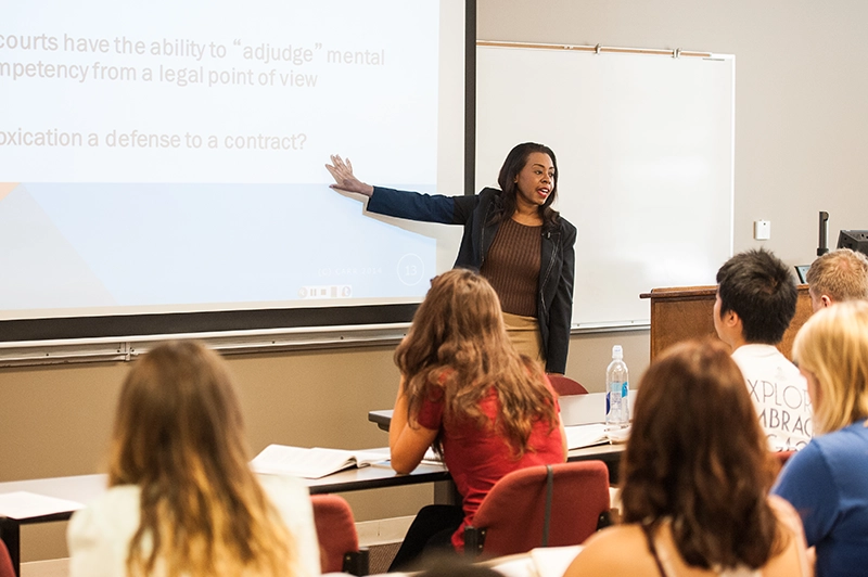 A female teacher at the front of a classroom filled with students gesturing to a projection on a screen with information.