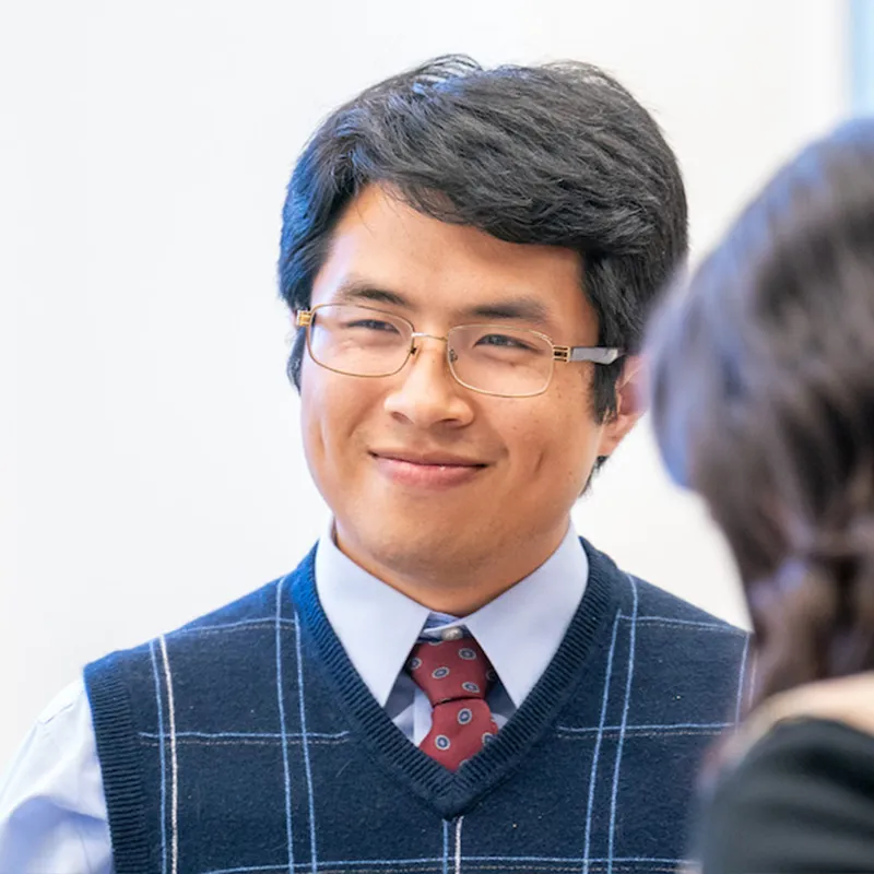 A student, dressed in a formal sweater vest, tie, and glasses, smiles during a conversation. His calm and pleasant demeanor suggests an engaging interaction in an academic or professional setting.