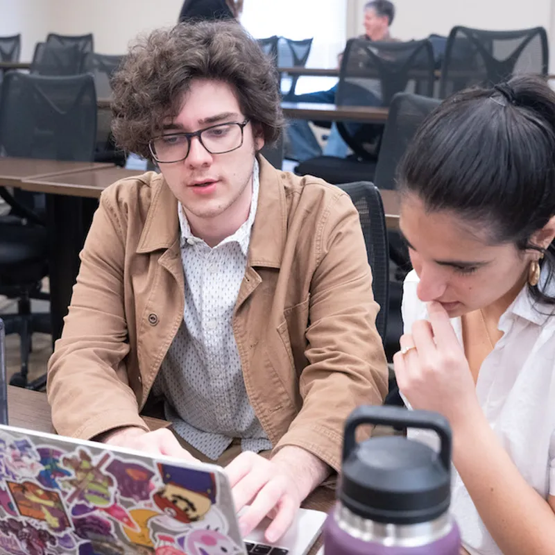 Two students collaborate on a project in a classroom. One student, wearing glasses and a brown jacket, is focused on his laptop covered with colorful stickers, while the other looks thoughtfully at the screen. The environment suggests a quiet study or research session.