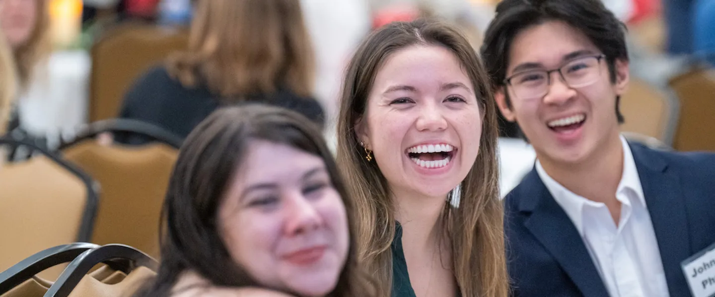 Three students smile and laugh while seated together at an event. The central student, a woman, has a wide, joyful smile, while the other two students, one male and one female, also appear happy and engaged. The setting is a formal or academic gathering, with chairs and tables visible in the background. The atmosphere is lively and social, reflecting a sense of camaraderie and connection, likely during a Global Honors program event at Belmont University.