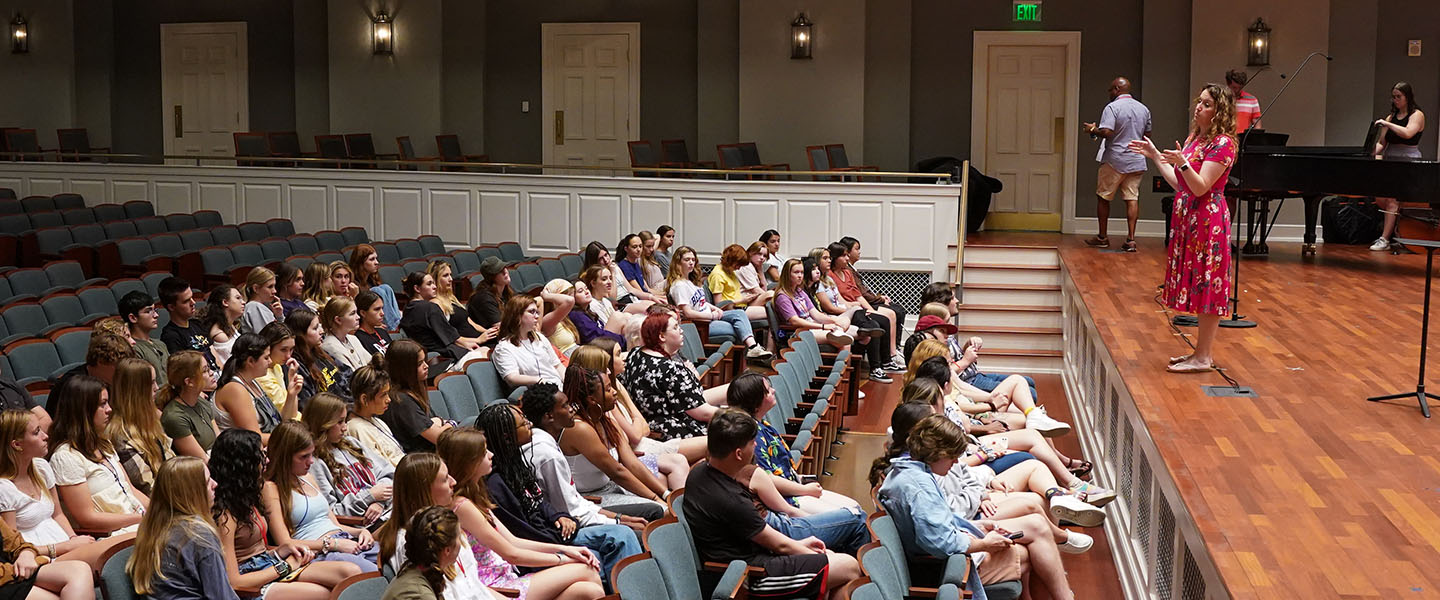 A group of students sit in theater seating listening to their professor teach from on stage.