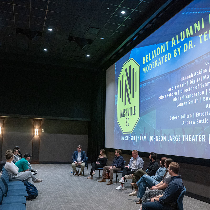 A group of panelists, seated in a semi-circle at the front, engage in conversation while an audience listens. The large screen behind them displays the Nashville SC logo and details of the event, which is moderated by Dr. Ted.