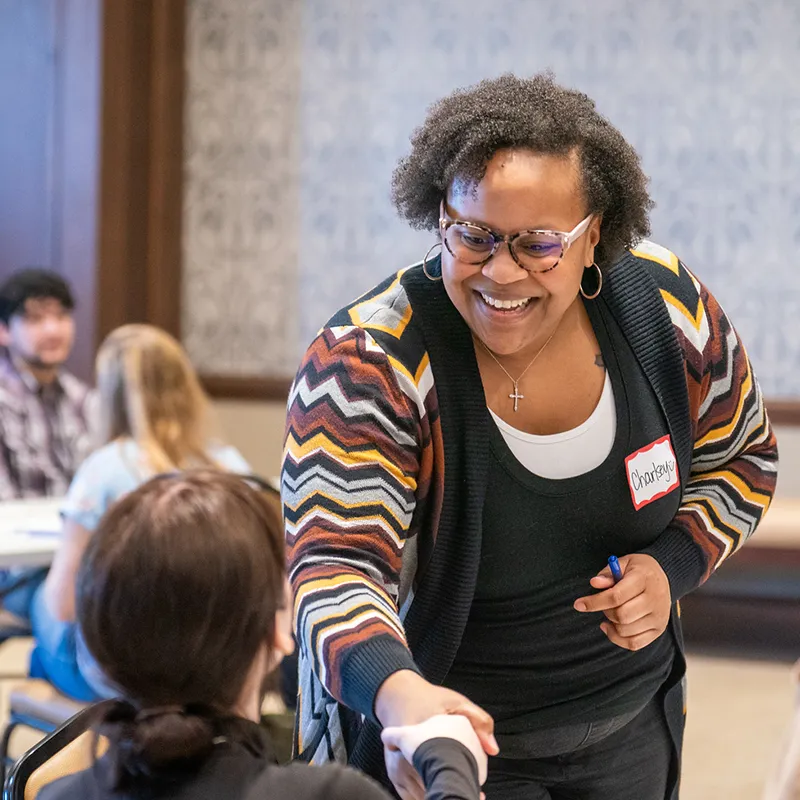 A smiling woman wearing glasses and a name tag interacts with another person during a social work event, with other attendees seated at tables in the background.