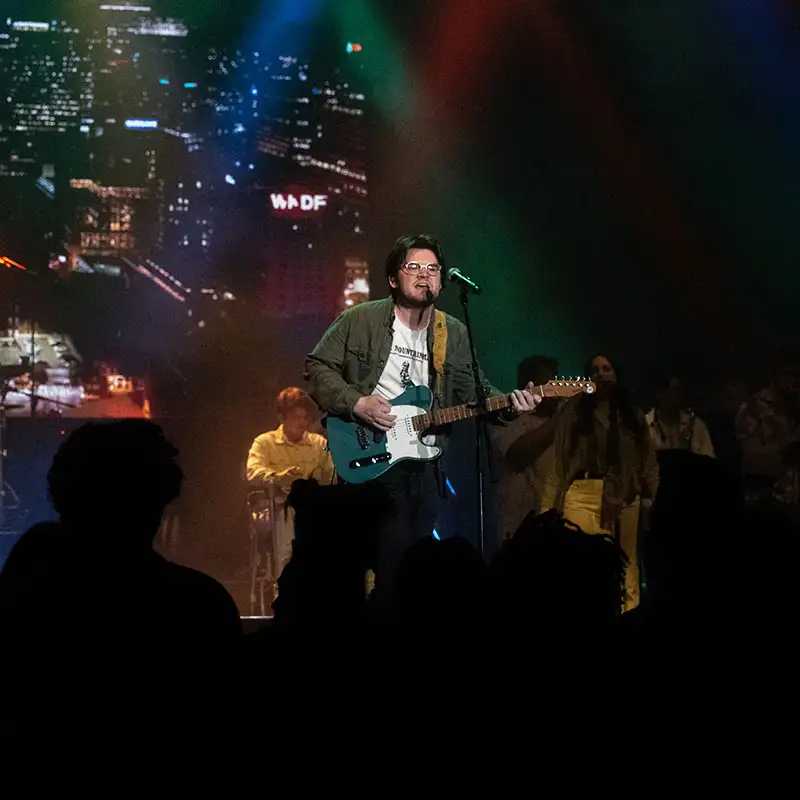 A musician performs on stage with a guitar, accompanied by a band, with colorful lights and a cityscape backdrop.