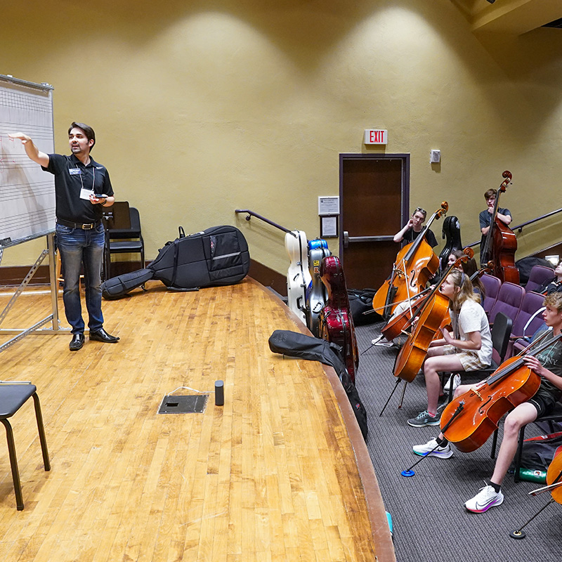 A teacher stands on stage and points to a white board while students sit in theater seating listening.