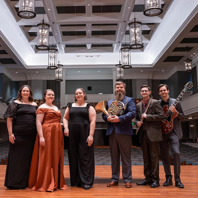 Group of musicians standing on a stage in a concert hall, dressed in formal attire, holding various instruments including a French horn, flute, and electric guitar, smiling for a group photo under elegant chandeliers.