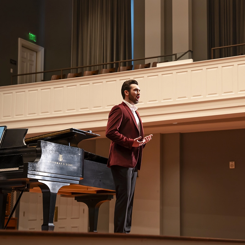 A vocalist performs passionately on stage beside a Steinway & Sons grand piano in an elegant concert hall.