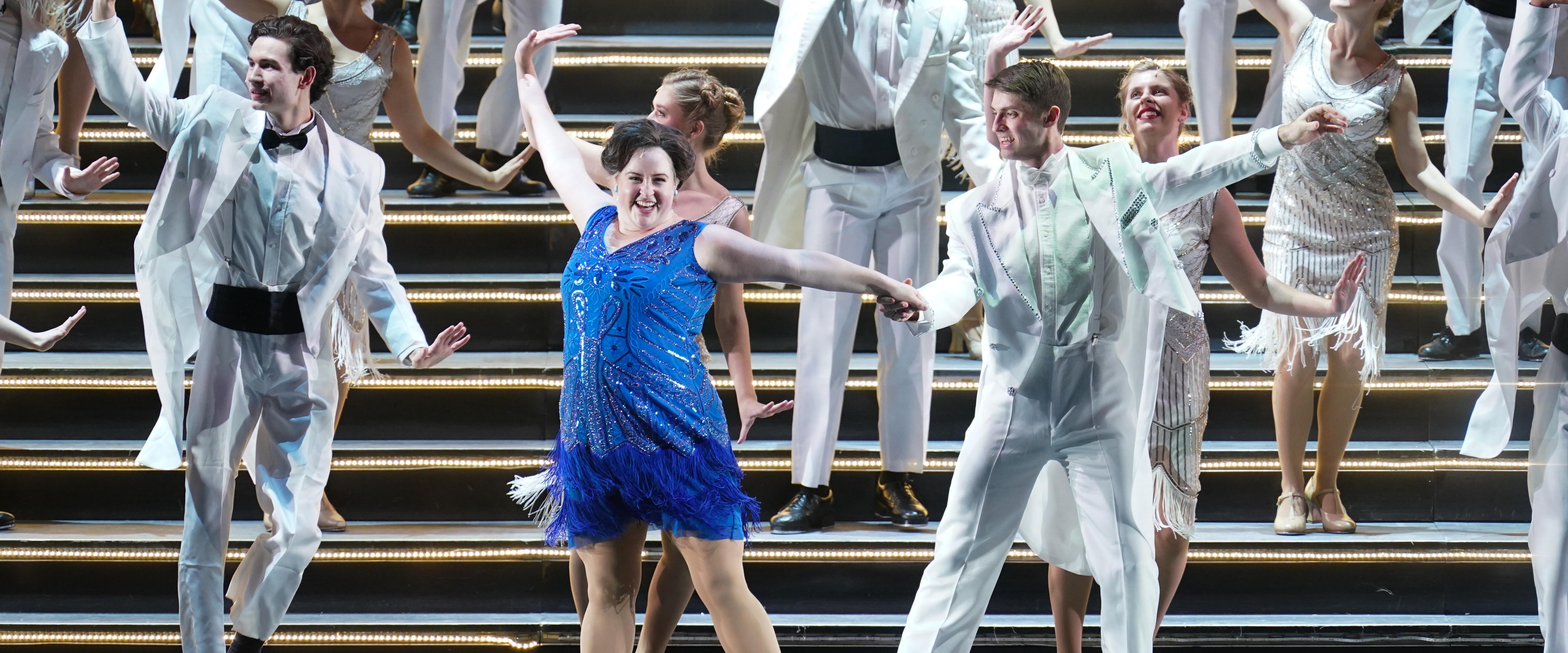 Two students sing with their arms raised on a stage lit by a border of bulb lights.