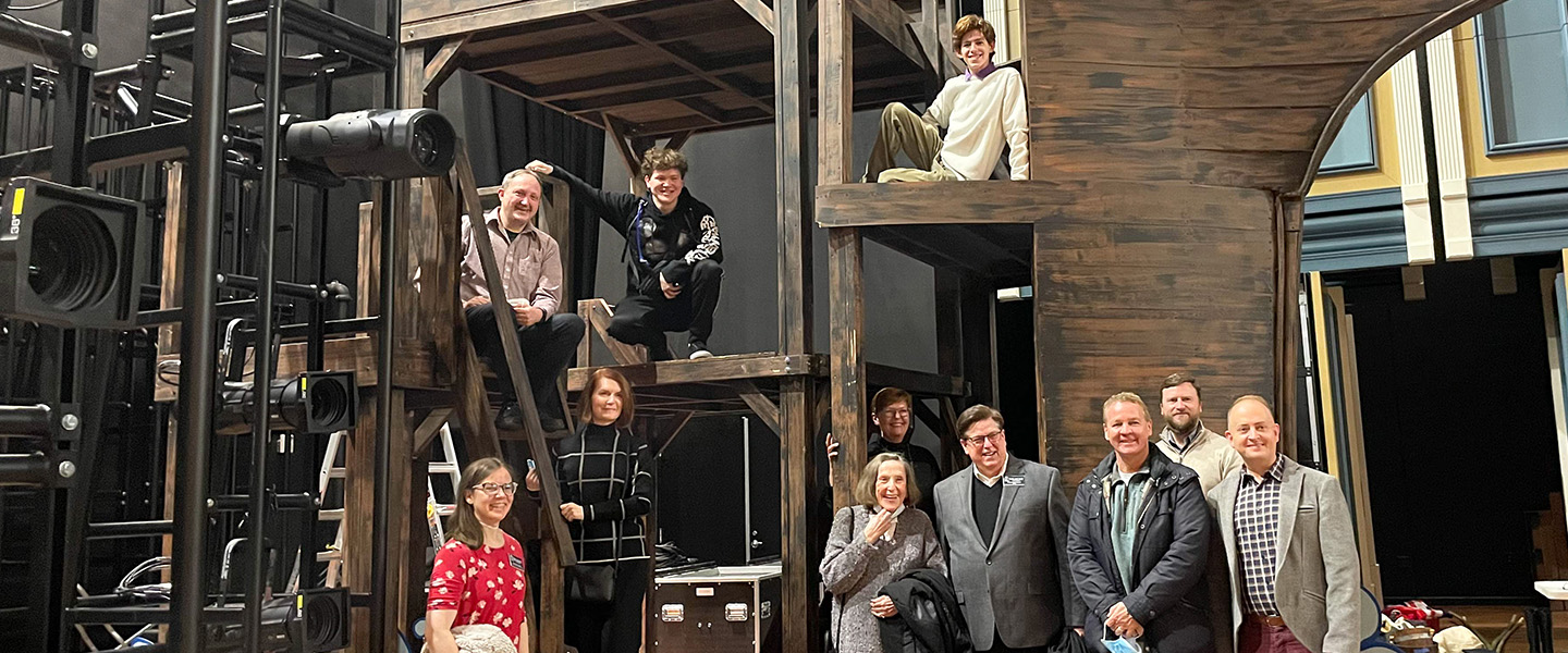 A group of students and staff stand in front of a set piece on stage at the Fisher Center.