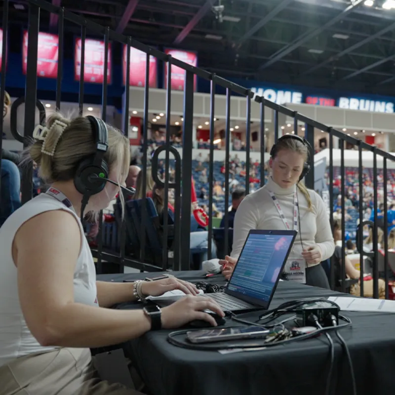 Two individuals working at a sports event in an arena. One is seated at a table with a laptop and headset, while the other is standing nearby, wearing a credential lanyard and headset. In the background, a crowd fills the arena, with 'Home of the Bruins' displayed prominently on the wall.