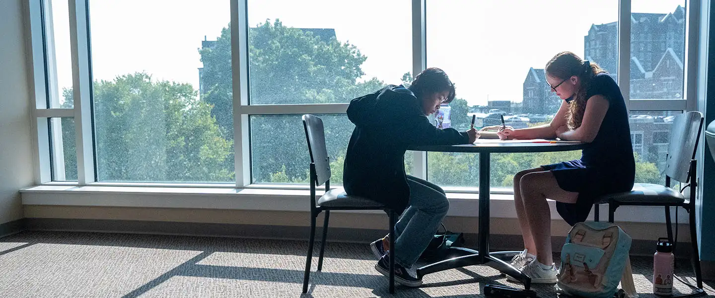 Two students working at a table in front of a window with a residence hall in the background