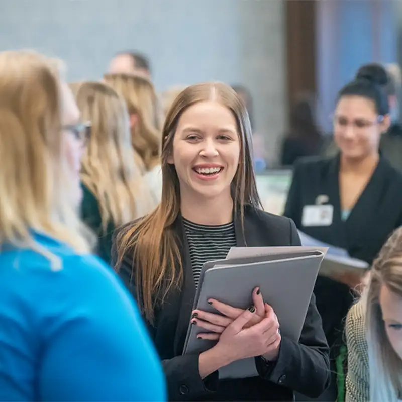 An indoor photo of a smiling woman dressed in business professional and holding a leather folio