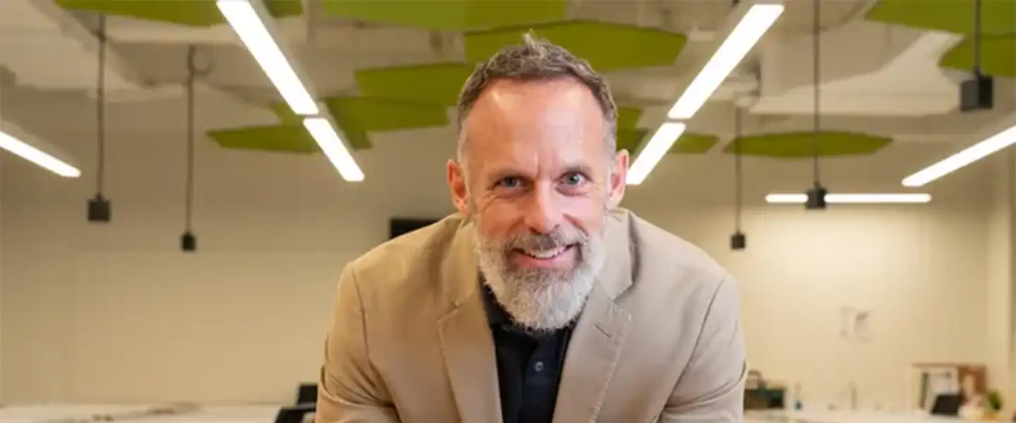 An indoor photo of a man smiling, leaning forward on a high table