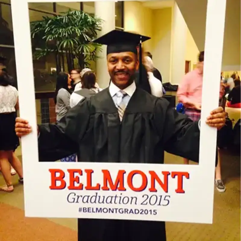 An indoor photo of an Adult Student in graduation cap and gown, holding a picture frame in front of him that reads "Belmont Graduation 2015 #BelmontGrad2015"