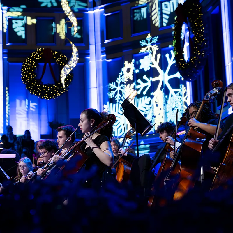 A symphony orchestra performs in a beautifully lit venue adorned with festive holiday decor, including wreaths, snowflake projections, and soft blue lighting. Musicians focus intently on their instruments, with cellists in the foreground.