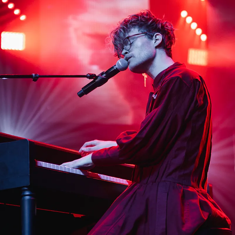 A musician plays the piano while singing into a microphone on stage. The performance is set against a dramatic red backdrop, with vibrant stage lighting illuminating the artist.