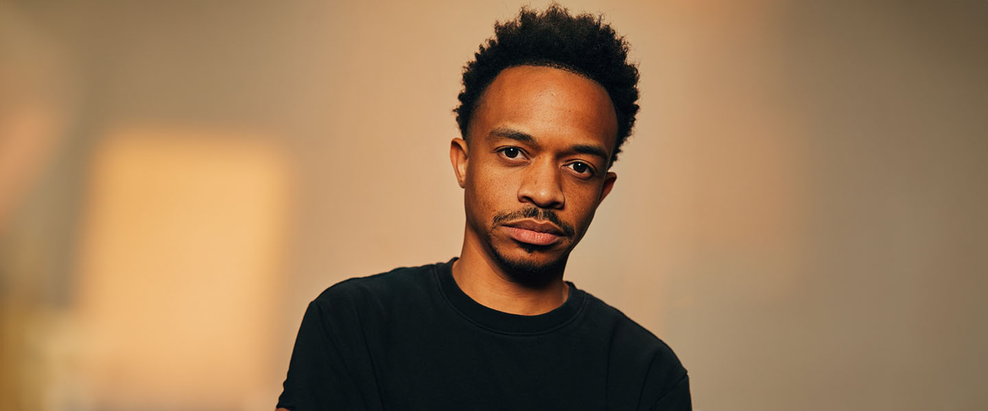 Dwan Hill poses against a neutral, softly lit background, creating a warm and focused atmosphere. He wears a casual black shirt and looks directly at the camera with a thoughtful, introspective expression.