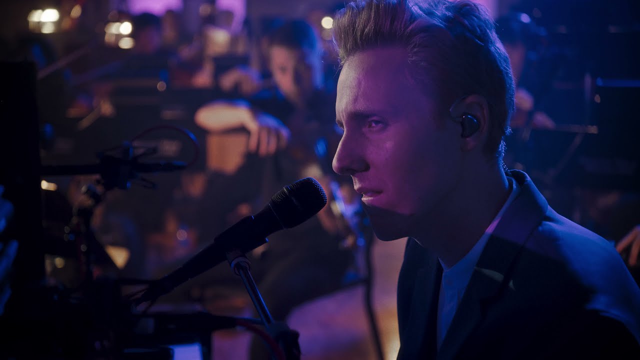 Cody Fry, illuminated by soft blue and purple stage lighting, sings into a microphone during a live performance. He is wearing an earpiece and is deeply focused, with an orchestra visible in the background, creating a dramatic and immersive musical atmosphere. The blurred figures of musicians emphasize Fry as the central performer. The scene exudes intensity and emotion, capturing the live concert's energy and Cody Fry's connection to the music.