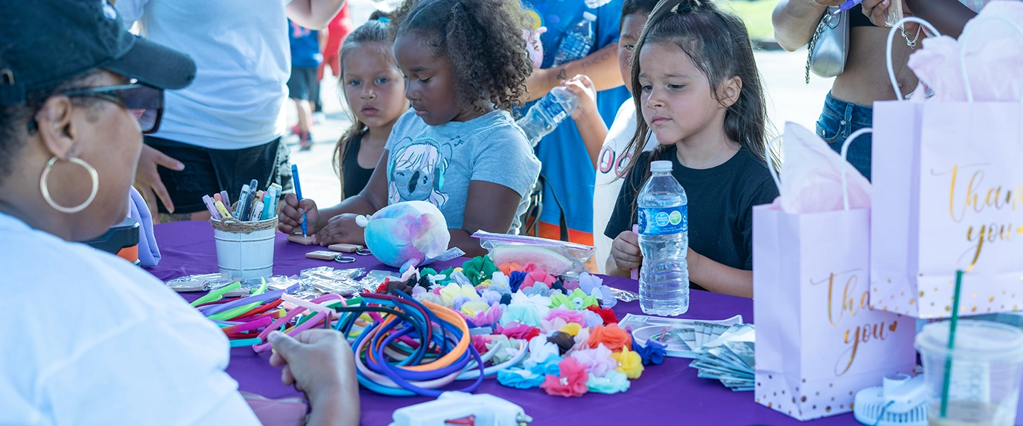 Taken at a community event, children at a table outdoors with brightly colored string and craft materials