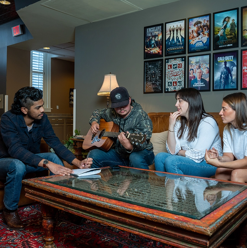 Students, one with a guitar, and another with a pen and paper are sitting on a leather couch, writing songs