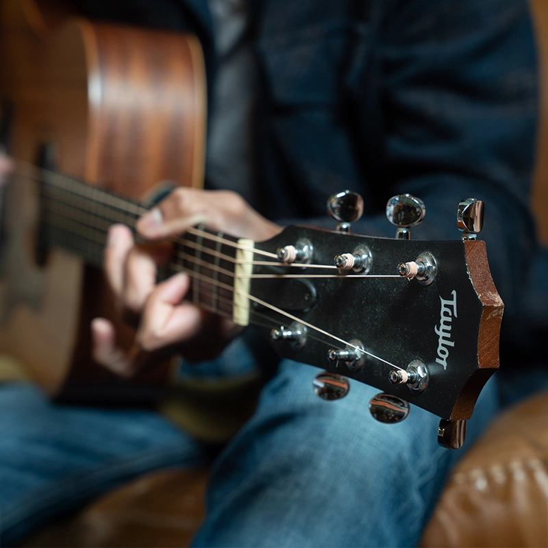 A close up of a Taylor guitar that is positioned on someone's lap in jeans