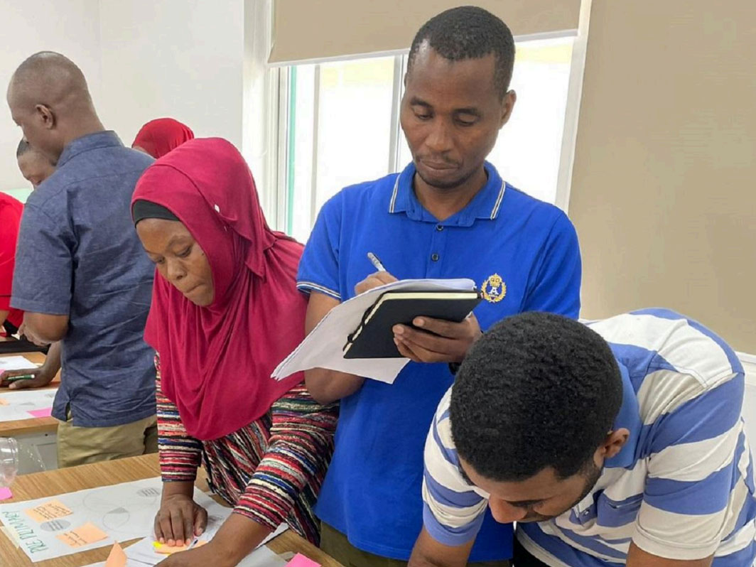 Group of participants working together during a workshop, with one person writing in a notebook and others collaborating at a table.