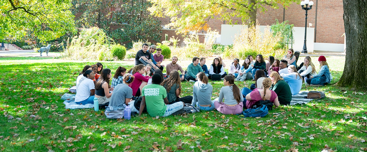 A class of students meeting outside and sitting on the grass at Belmont during a sunny day