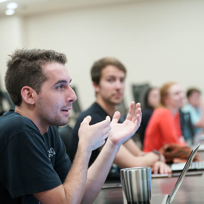 Student speaking in class at table with his laptop open in front of him