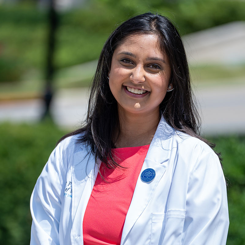 A smiling student from the inaugural class of the Thomas F. Frist, Jr. College of Medicine at Belmont University is wearing a white lab coat and standing outdoors. She has long dark hair and is wearing a bright coral dress underneath her coat. 