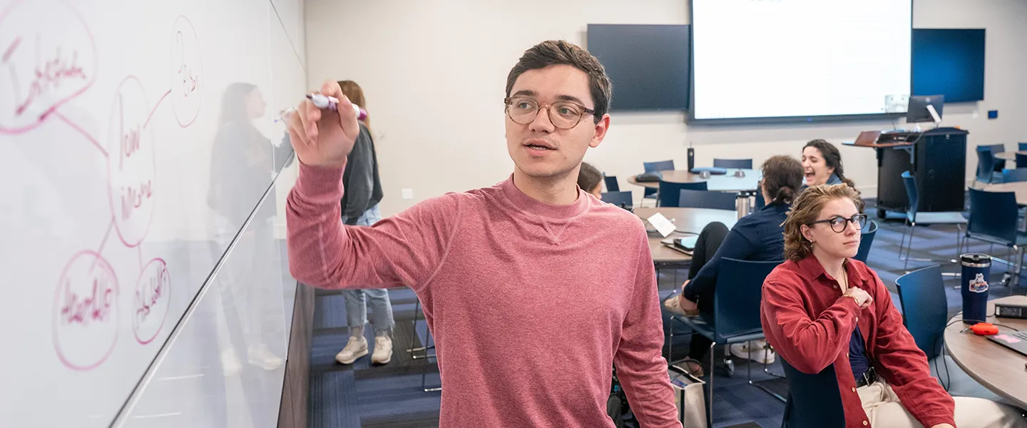 A student wearing glasses and a red sweater stands at a whiteboard in a classroom, writing a mind map with interconnected ideas in red marker. He appears focused and engaged in his task. In the background, other students are seated at tables, collaborating and discussing, with one student in a red jacket turning towards the camera. The classroom is modern, with large screens and collaborative seating arrangements. The scene reflects an active, interactive learning environment.