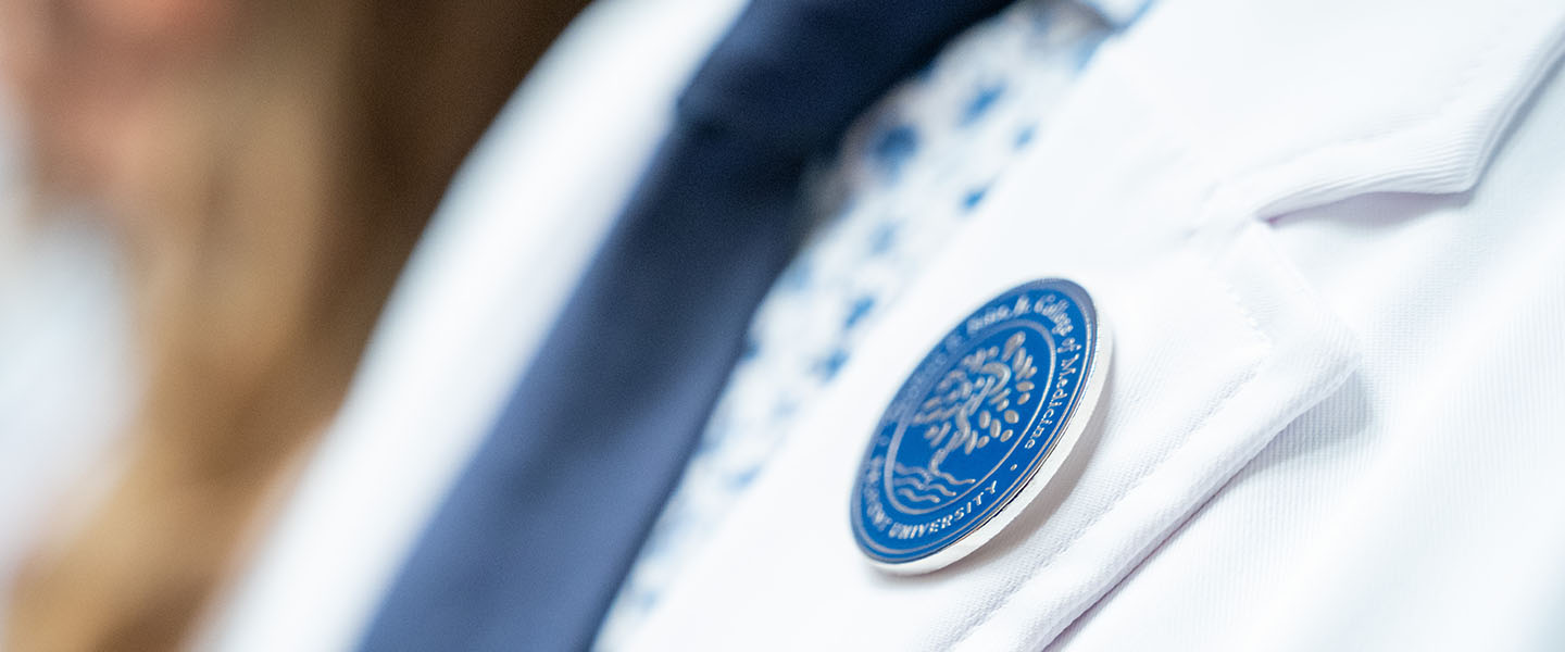 Close-up of a Belmont University medical student's white coat, focusing on a circular blue and white seal of the Thomas F. Frist, Jr. College of Medicine pinned to the coat.