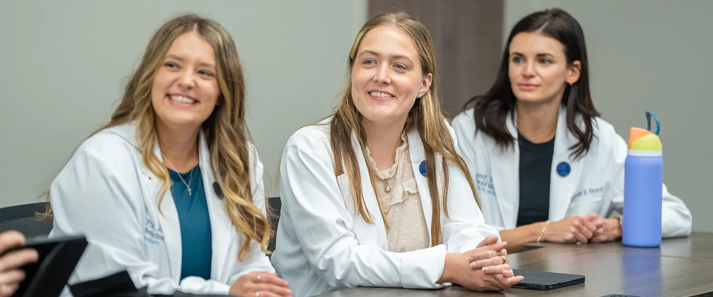 Three medical students are seated at a table, smiling and engaged in conversation. They are all wearing white lab coats, with one student in the middle laughing while interacting with the group. A brightly colored water bottle sits on the table near the student on the right. The setting appears to be a classroom or meeting room, reflecting a collaborative and positive learning environment.