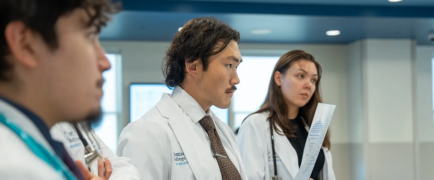 A group of medical students from the Frist College of Medicine at Belmont University are shown in a clinical setting. They are wearing white lab coats and are focused on a discussion. One student in the foreground holds a piece of paper, likely reviewing medical notes, while listening intently. The background features a modern, well-lit environment, suggesting an educational or healthcare setting.