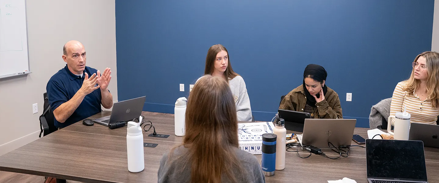 A group of students and a professor are seated around a table in a classroom, engaged in discussion. The professor, sitting at the head of the table, is speaking and gesturing with his hands. The students, equipped with laptops and notebooks, appear attentive, with one student taking notes while others listen or work on their computers. The setting is a modern classroom with a blue accent wall, whiteboard, and minimal distractions, indicating a focused learning environment. Several water bottles are present, reflecting a casual and collaborative atmosphere.