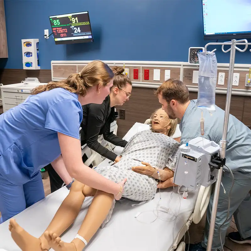  Three healthcare professionals are attending to a patient simulation doll in a hospital room. One nurse in scrubs is adjusting the doll's position, while another nurse in a black shirt and a male nurse in scrubs are monitoring the doll's medical equipment. A monitor displaying vital signs is visible in the background.