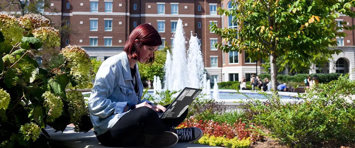 An outdoor photo on a sunny day of a female student seated with a laptop on her lap on Belmont's campus