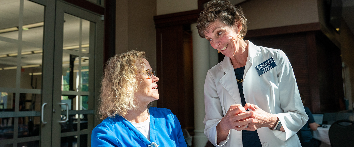 A medical professional in a white lab coat smiling and speaking with someone in blue scrubs with the sun highlighting their faces.