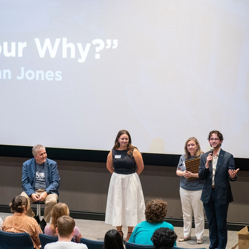 Garrett Jacob holds a microphone and gestures while addressing an audience in a lecture hall. Beside him, Lauren Prickett smiles, while another woman holds a wooden plaque. President Greg Jones observes. A large screen behind them displays the words “What’s Your Why?” as part of a presentation. Audience members are visible in the foreground.