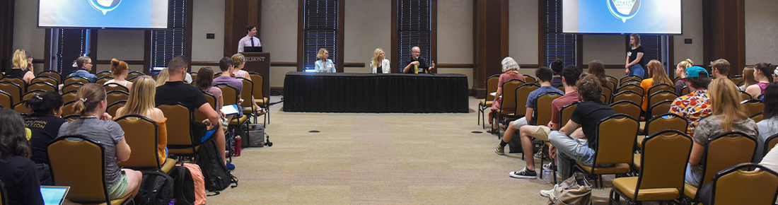 A panel of speakers and a moderator seated in the front of an open room speaking and presenting to an audience