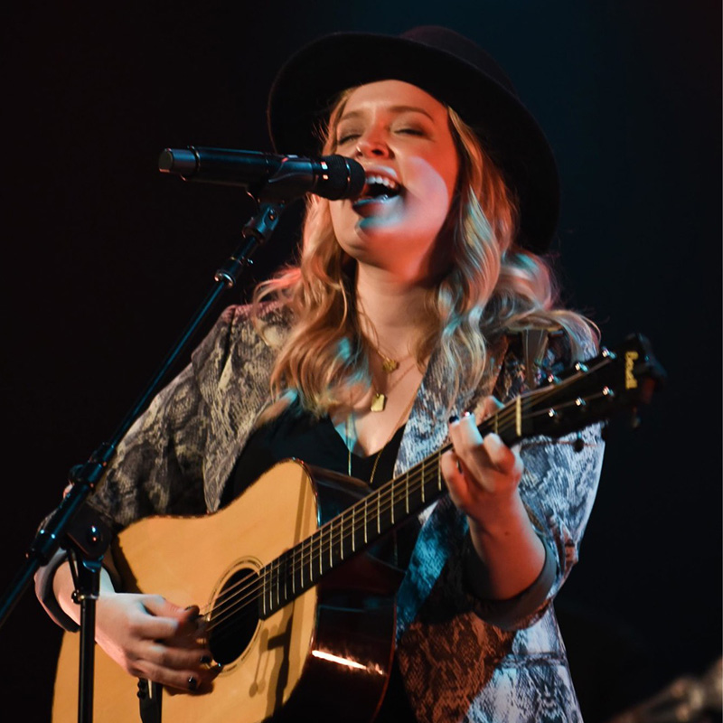 Student performing on stage, passionately singing into a microphone while playing an acoustic guitar.