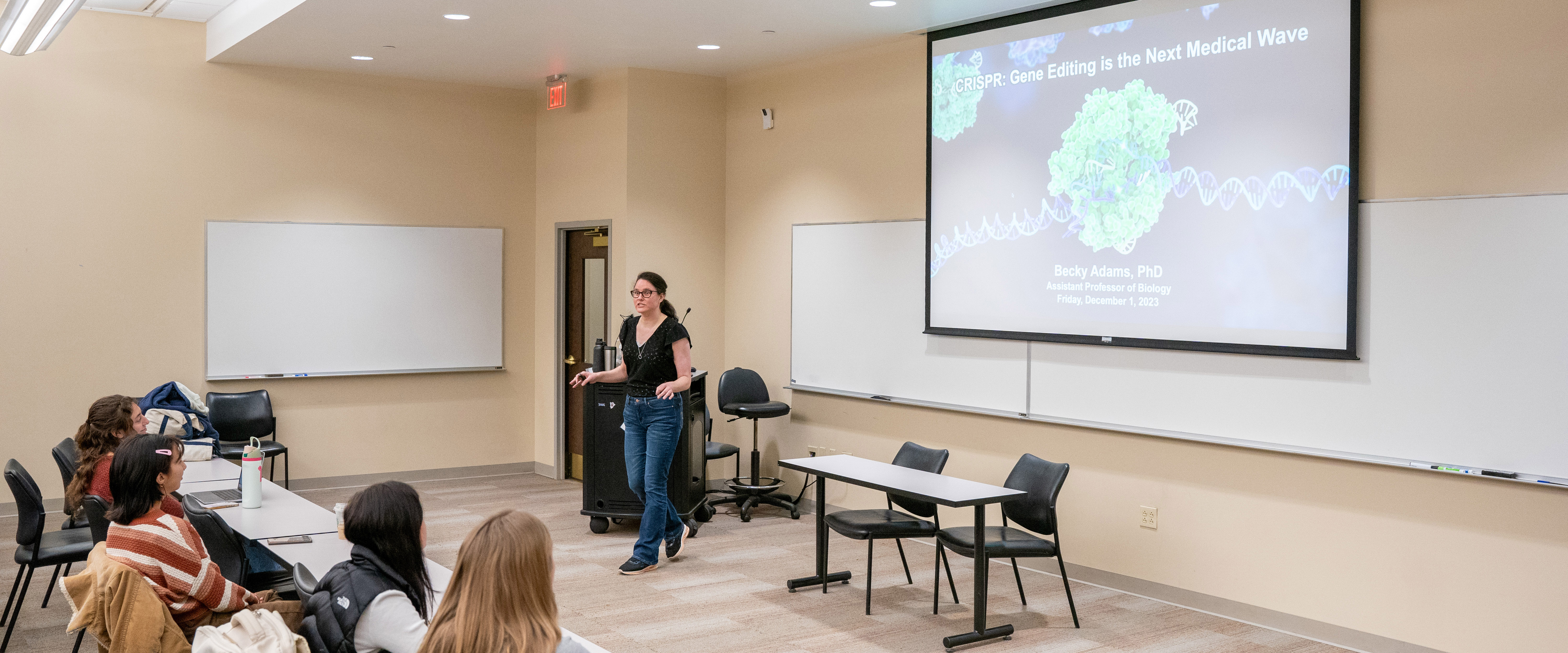 Science professor talking to class of students with a presentation behind her. 