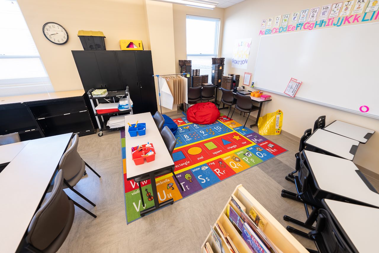 Elementary classroom interior with colorful educational decor. Features include alphabet letters on the wall, a number and shape floor rug, desks, chairs, storage cabinets, and various learning materials. The room has a bright and inviting atmosphere.