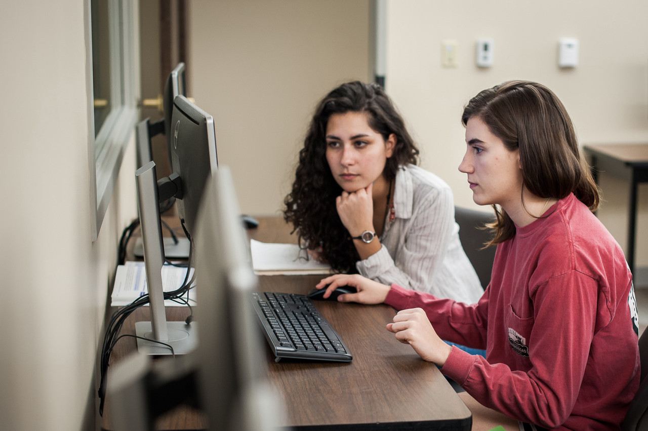 Two students work together at a desktop computer in a classroom or computer lab. Both are focused on the screen, with one student operating the mouse while the other observes closely. The workspace is equipped with multiple computer stations, indicating a shared learning or collaborative environment. The students' serious expressions suggest they are engaged in problem-solving or completing an academic project. The setting is a modern, organized lab, providing a quiet place for concentration and teamwork.