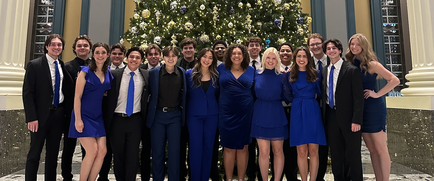 Calista Morrison in blue power suit with vocal ensemble in front of a Christmas tree