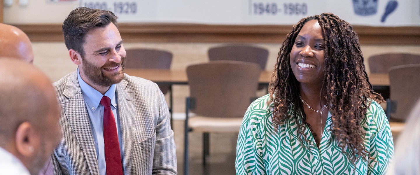 Two professionals in a meeting or conference room setting sharing a genuine laugh or moment of joy. One person is wearing a beige suit jacket with a red tie, and the other is wearing a green and white patterned dress or top.