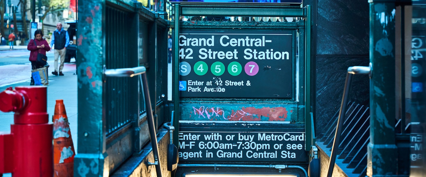 A New York City subway entrance sign for Grand Central-42 Street Station, featuring colored circles for lines S, 4, 5, 6, and 7. The sign includes accessibility information and operating hours (M-F 6:00am-7:30pm). The sign appears weathered with some graffiti, and has a blue-teal tint from the surrounding lighting. Above the entrance sign is a digital display showing "No real time data Listen for announcements"